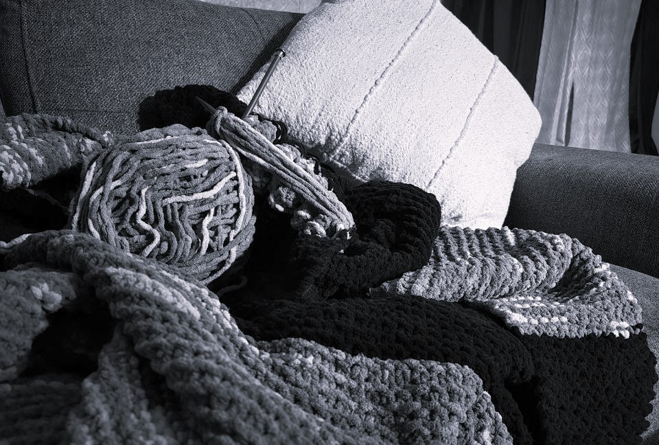 An in-progress knitted blanket draped across a couch in black and white, yarn ball and needle resting mid-row